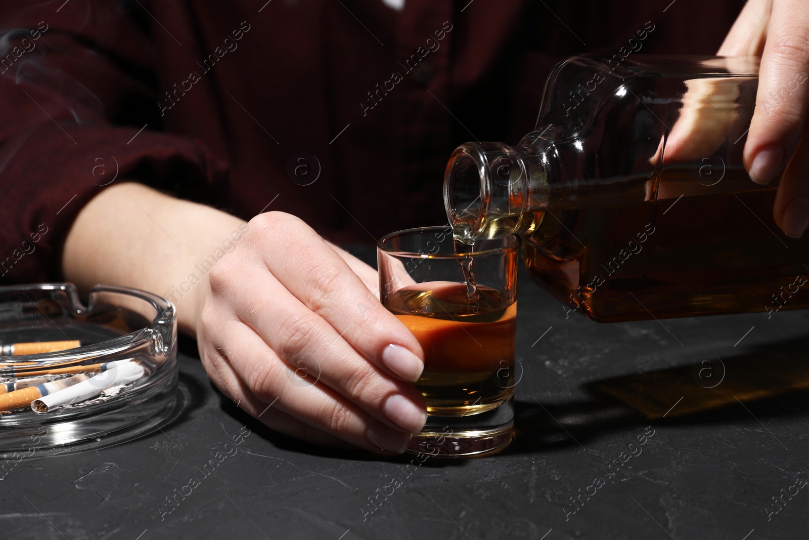 Photo of Alcohol addiction. Woman pouring whiskey from bottle into glass at dark textured table, closeup