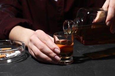 Alcohol addiction. Woman pouring whiskey from bottle into glass at dark textured table, closeup