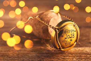 Cork of sparkling wine and muselet cap with engraving Happy 2023 New Year on wooden table, closeup. Bokeh effect