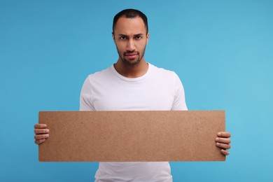 Photo of Upset man holding blank cardboard banner on light blue background, space for text