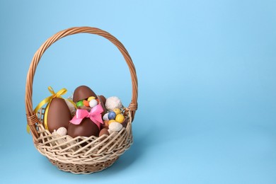 Photo of Wicker basket with tasty chocolate Easter eggs and different candies on light blue background, space for text