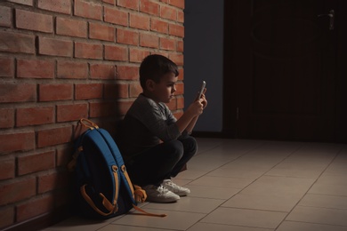 Sad little boy with mobile phone sitting on floor near brick wall indoors