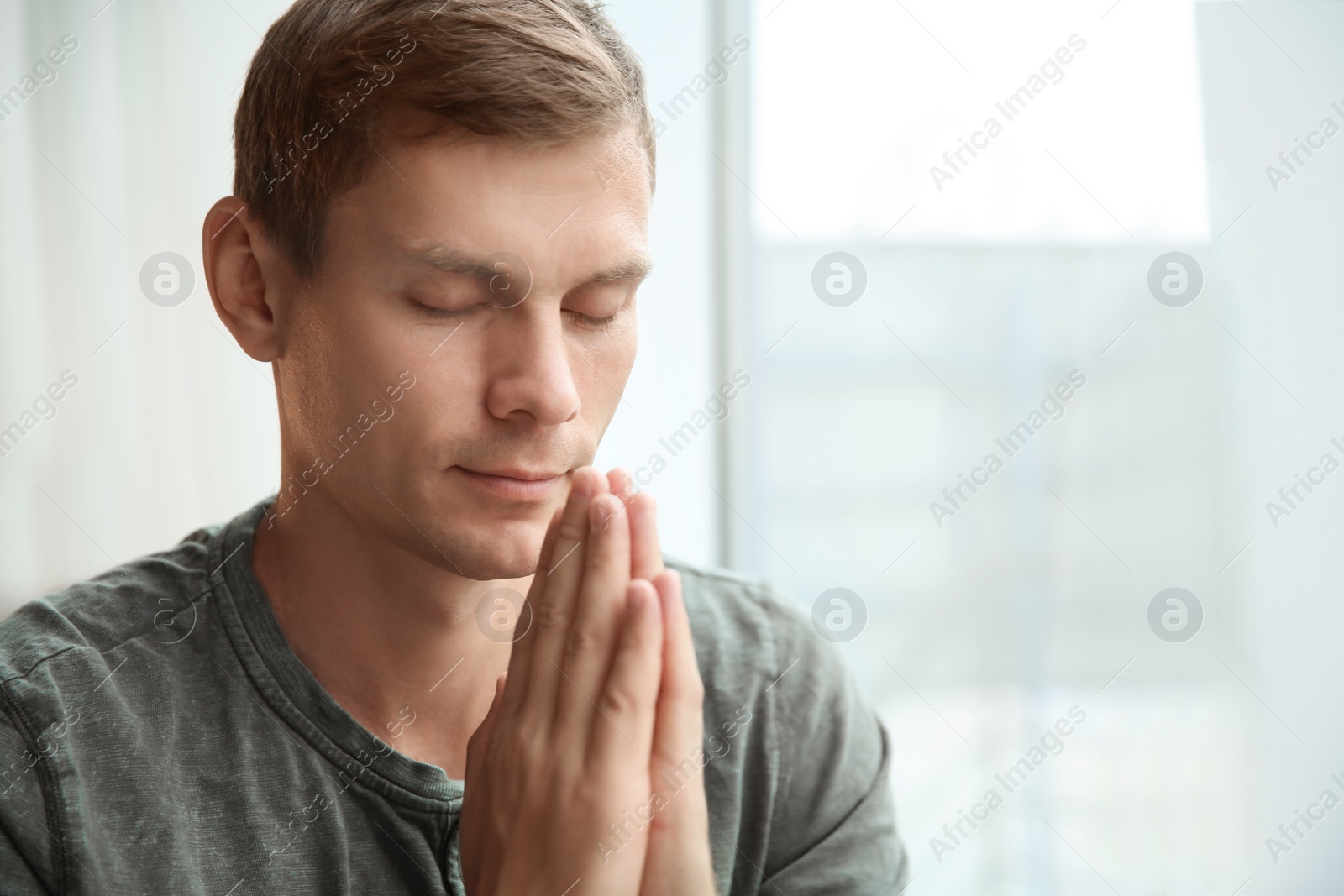 Photo of Man with hands clasped together for prayer near window. Space for text