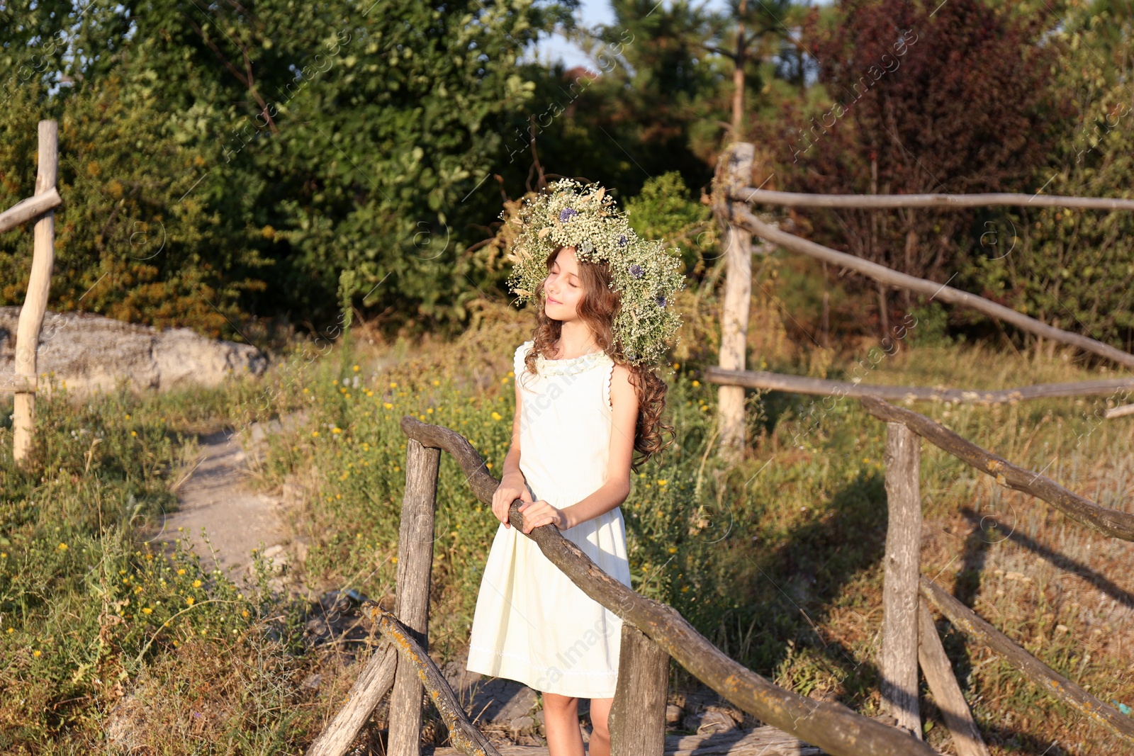 Photo of Cute little girl wearing wreath made of beautiful flowers on wooden bridge