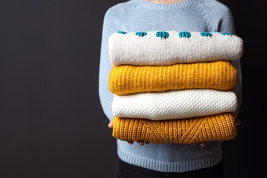 Woman holding pile of winter sweaters on dark background, closeup view. Space for text