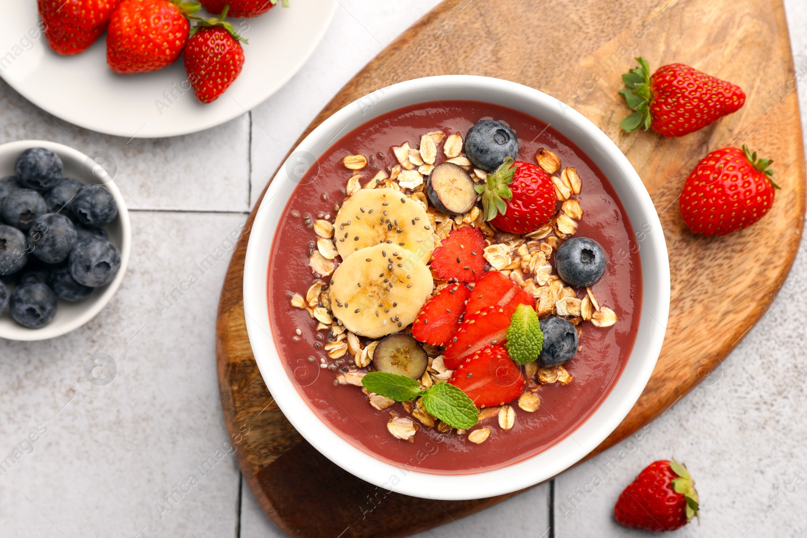 Photo of Delicious smoothie bowl with fresh berries, banana and granola on tiled surface, flat lay