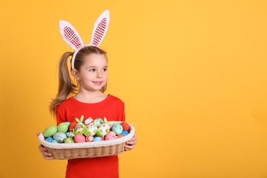 Photo of Adorable little girl with bunny ears holding wicker basket full of Easter eggs on orange background. Space for text
