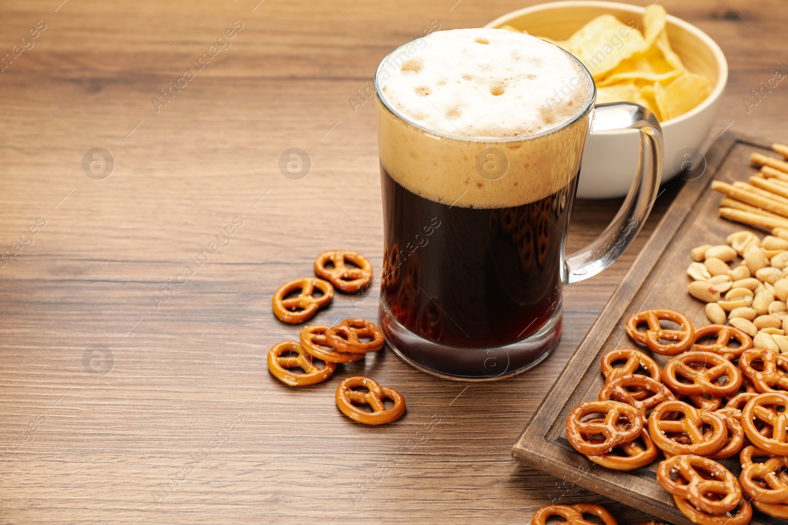 Photo of Glass of beer served with delicious pretzel crackers and other snacks on wooden table, space for text