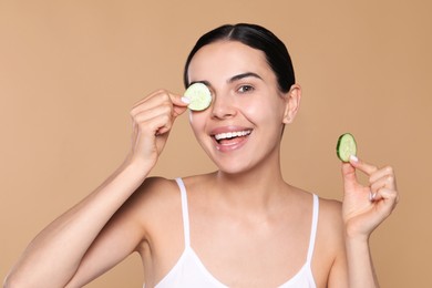 Photo of Beautiful young woman putting slices of cucumber on eyes against beige background