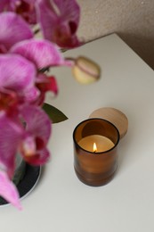 Photo of Burning candle in glass container near blooming orchid on white table
