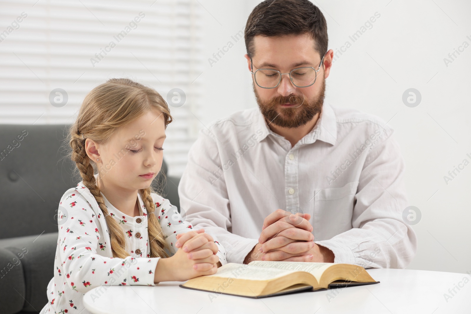 Photo of Girl and her godparent praying over Bible together at table indoors