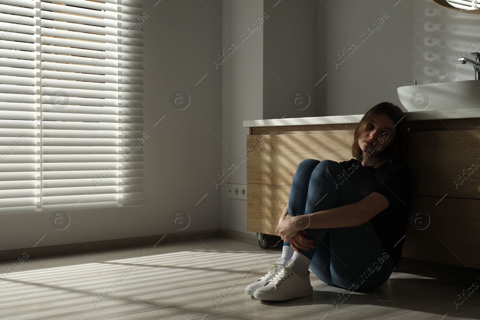 Photo of Sad young woman sitting on floor in bathroom, space for text