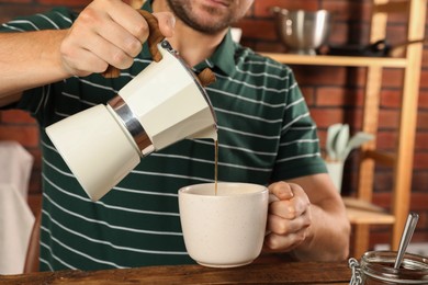 Photo of Man pouring aromatic coffee from moka pot into cup at table indoors, closeup