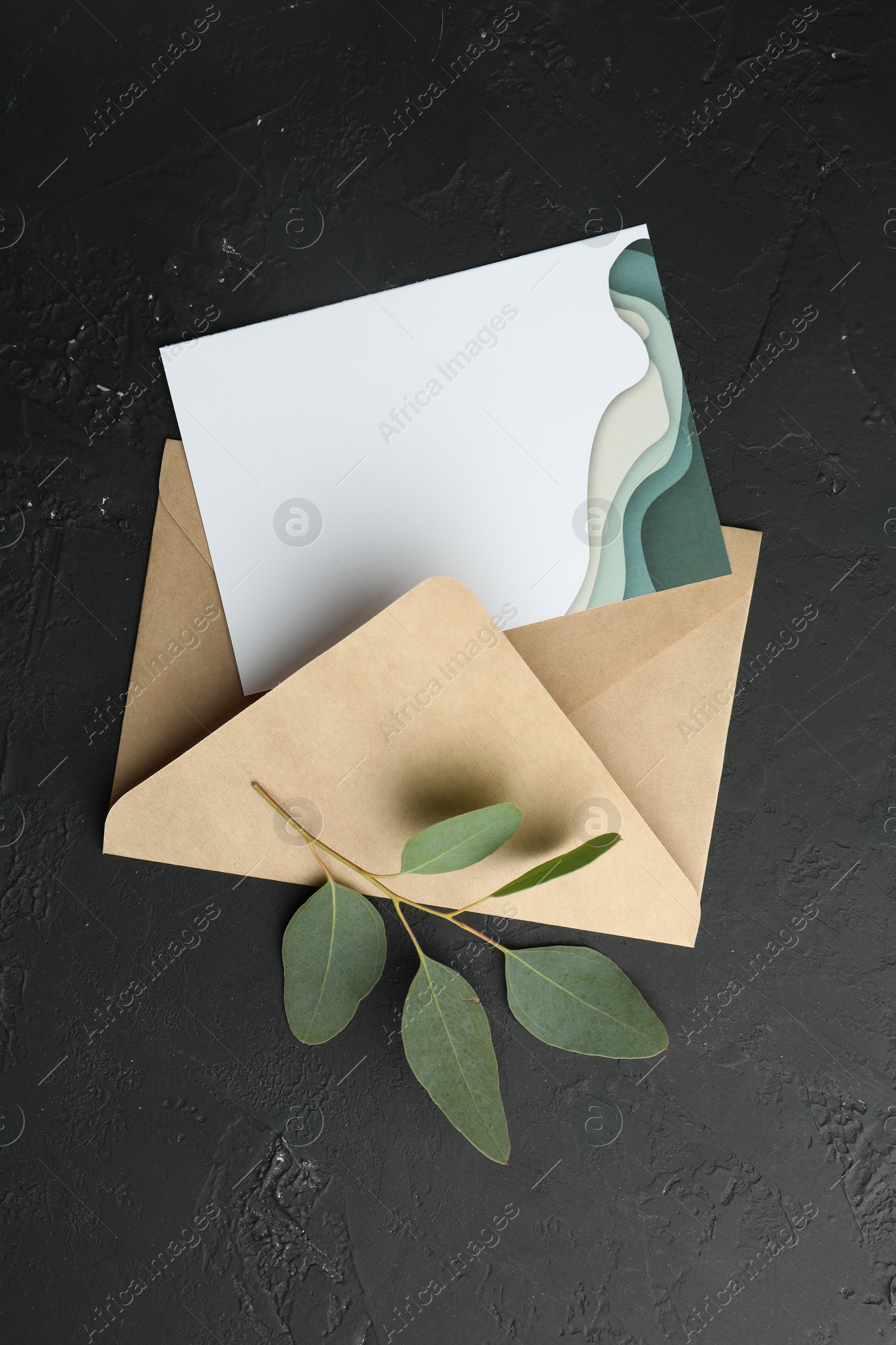 Photo of Envelope with blank invitation card and green leaves on black textured background, top view. Space for text