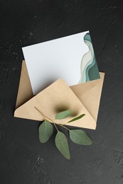 Envelope with blank invitation card and green leaves on black textured background, top view. Space for text