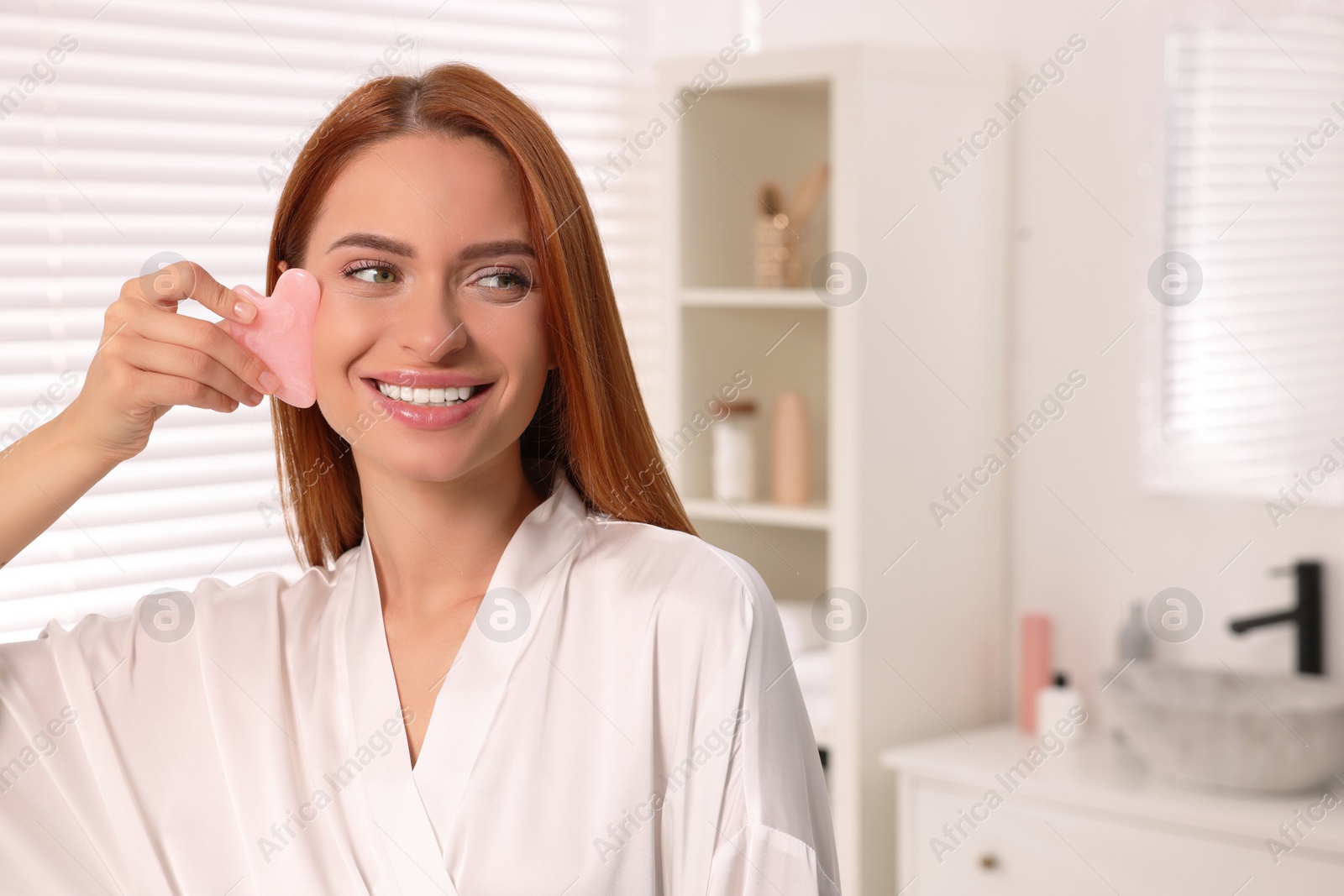 Photo of Young woman massaging her face with rose quartz gua sha tool in bathroom, space for text