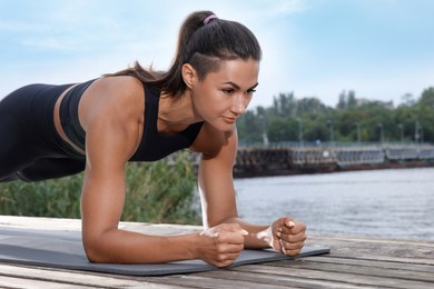 Photo of Young woman doing plank exercise on wooden pier near river