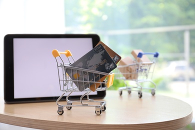 Photo of Internet shopping. Small cart with credit cards near modern tablet on table indoors