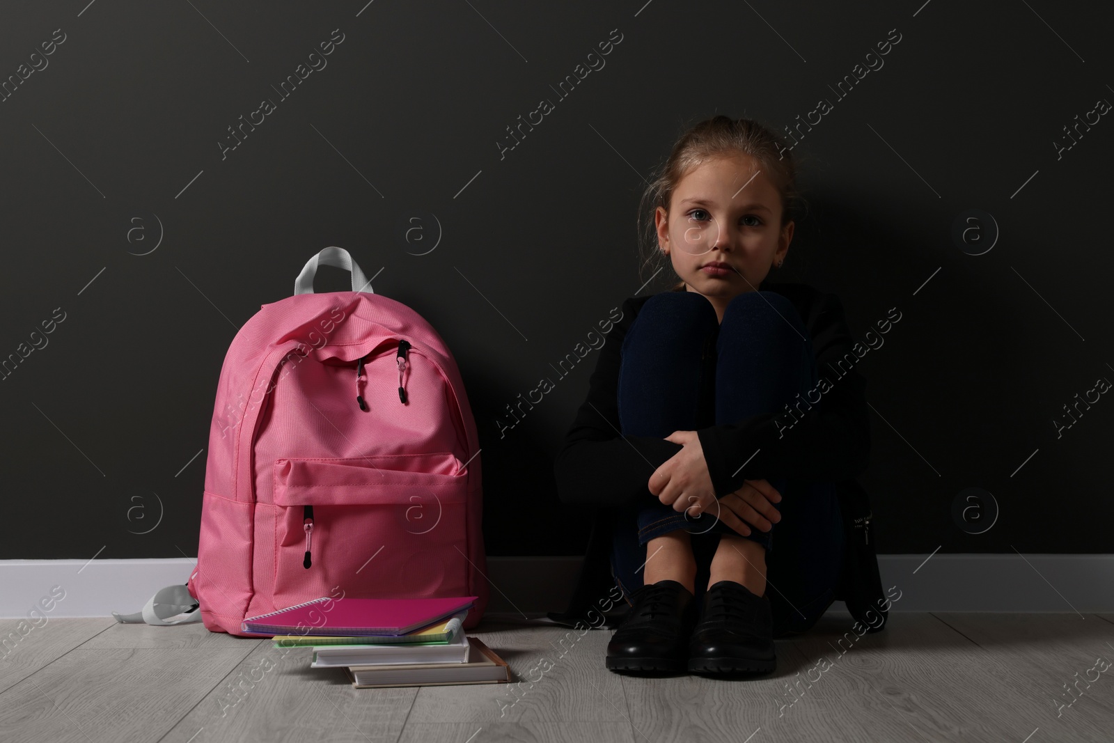 Photo of Upset girl with backpack sitting on floor near black wall. School bullying