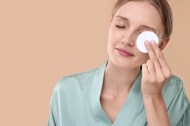 Young woman cleaning her face with cotton pad on beige background. Space for text