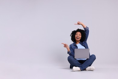 Happy young woman with laptop pointing at something on light grey background. Space for text