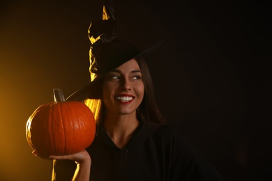 Young woman wearing witch costume with pumpkin on black background, space for text. Halloween party