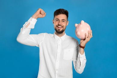 Happy young man with piggy bank on light blue background