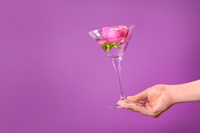 Photo of Woman holding martini glass with water and pink flowers on violet background, closeup. Space for text