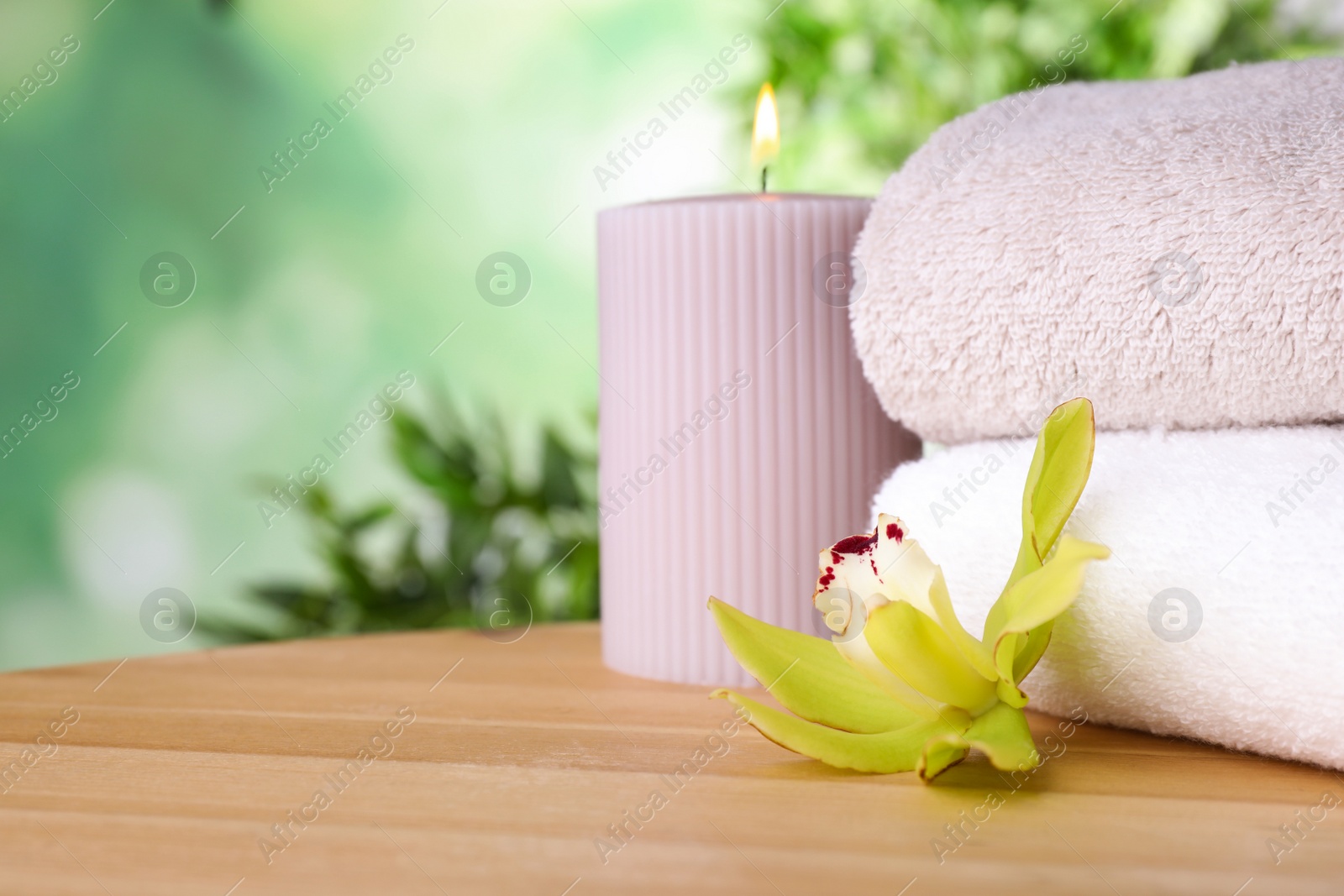 Photo of Exotic flower, burning candle and fresh towels on wooden table against blurred green background, space for text