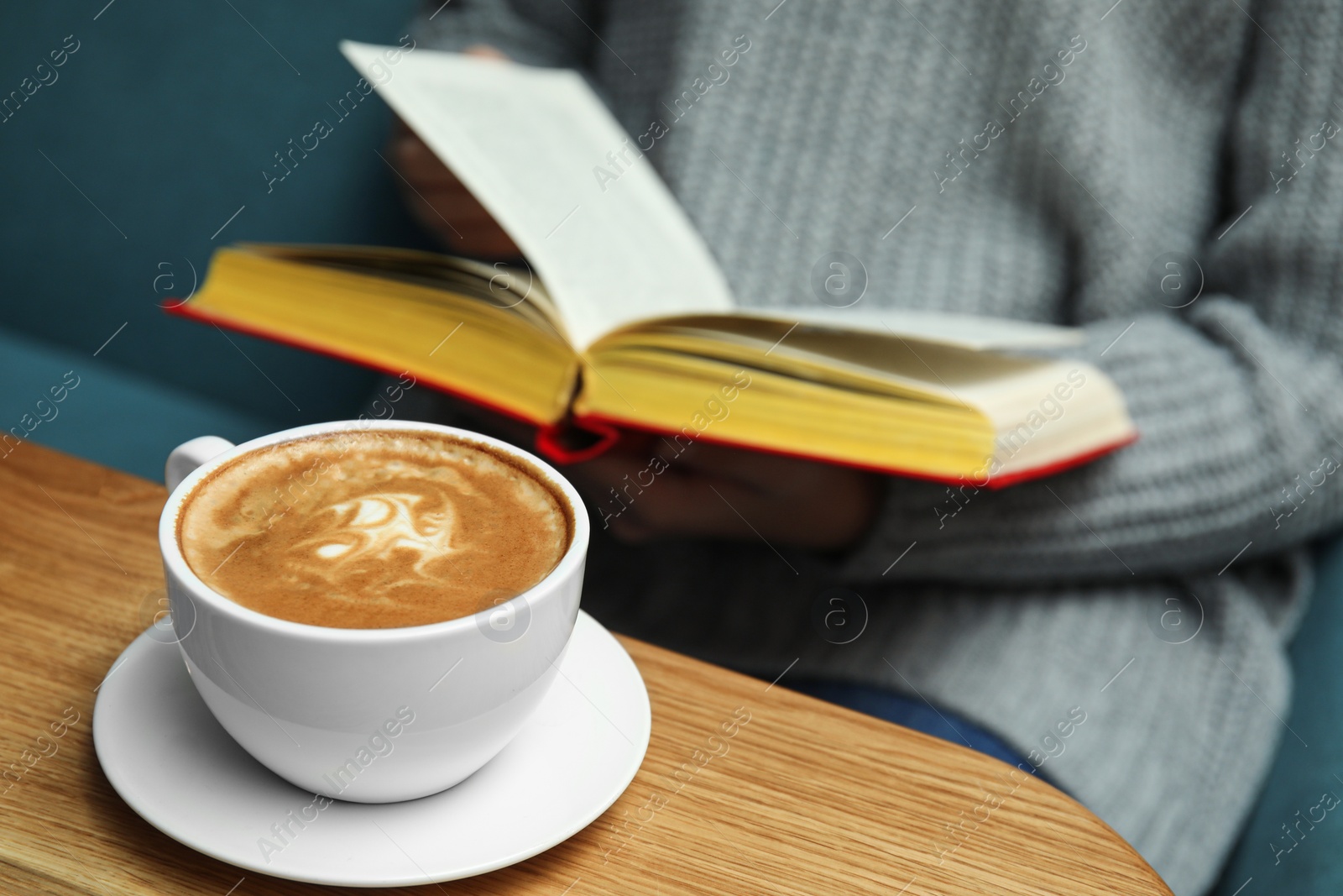 Photo of Woman reading book at table, focus on cup of coffee