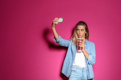 Beautiful woman with tasty drink taking selfie on color background
