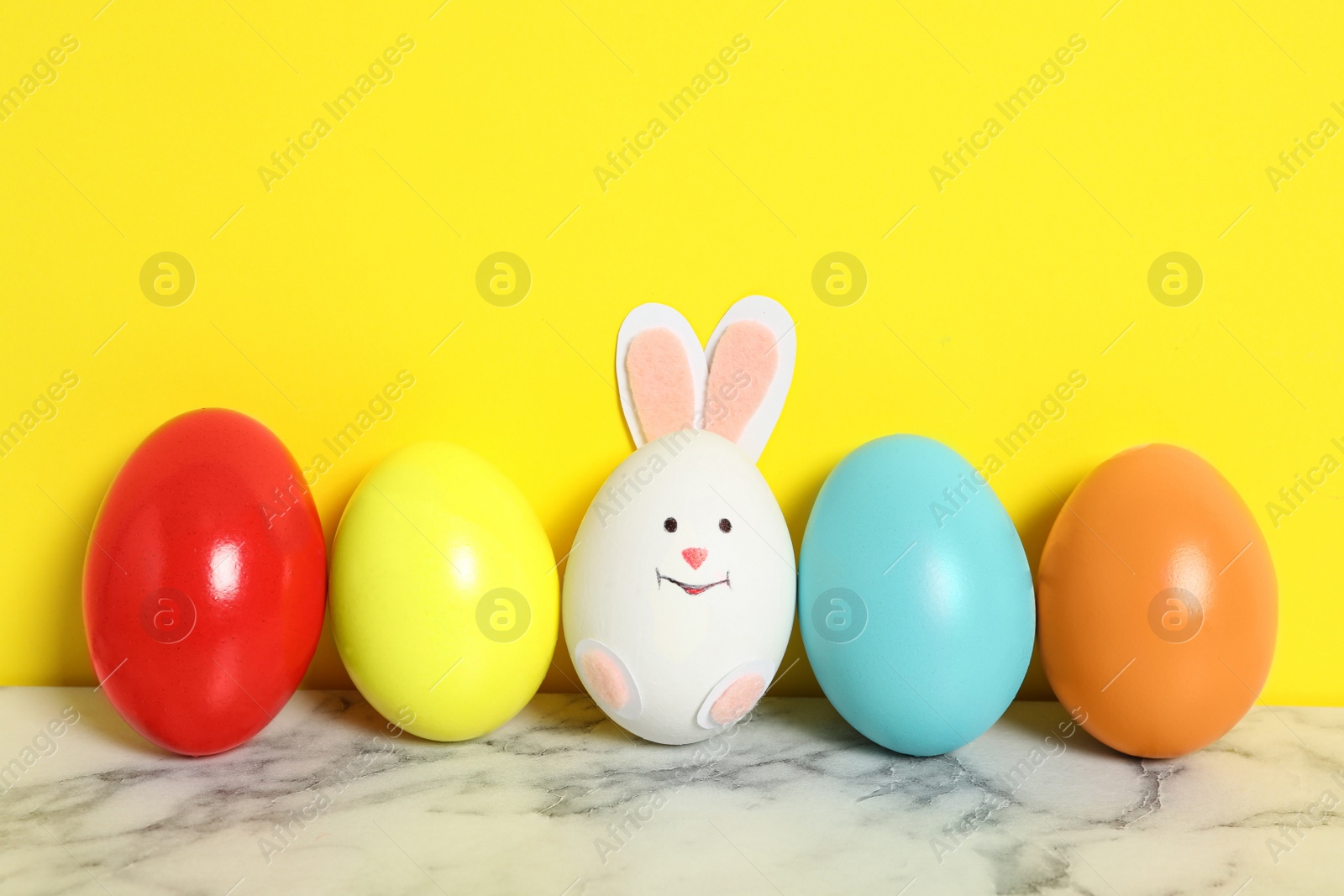 Photo of Bright eggs and white one as cute bunny on marble table against yellow background. Easter celebration