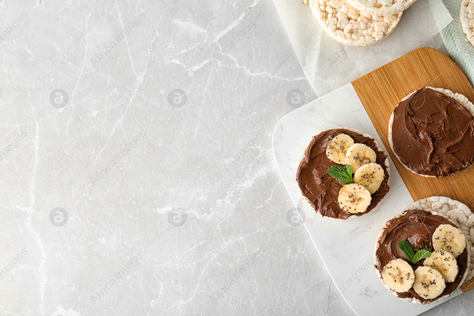 Photo of Puffed rice cakes with chocolate spread, banana and mint on grey marble table, flat lay. Space for text