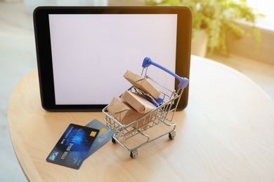 Internet shopping. Small cart with boxes and credit cards near modern tablet on table indoors