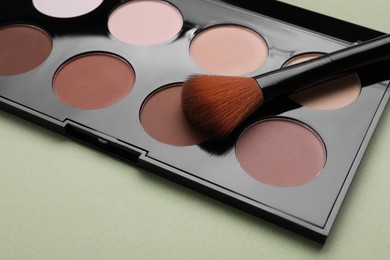 Photo of Colorful contouring palette with brush on pale green background, closeup. Professional cosmetic product
