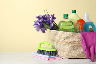 Spring cleaning. Wicker basket with detergents, flowers and gloves near tools on white wooden table. Space for text