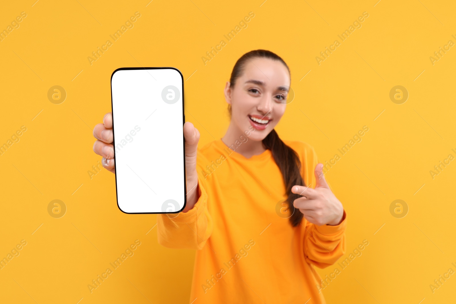 Photo of Young woman showing smartphone in hand and pointing at it on yellow background, selective focus. Mockup for design