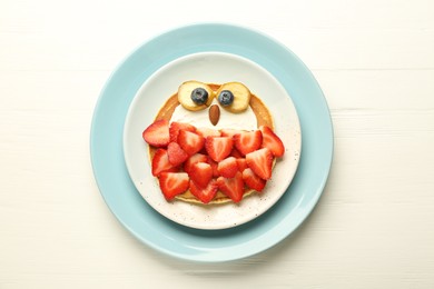 Photo of Creative serving for kids. Plate with cute owl made of pancakes, strawberries, cream, banana and almond on white wooden table, top view