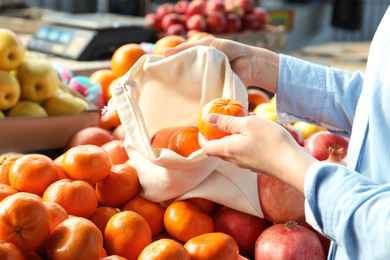 Photo of Woman putting tangerine into cotton eco bag at wholesale market, closeup. Life without plastic