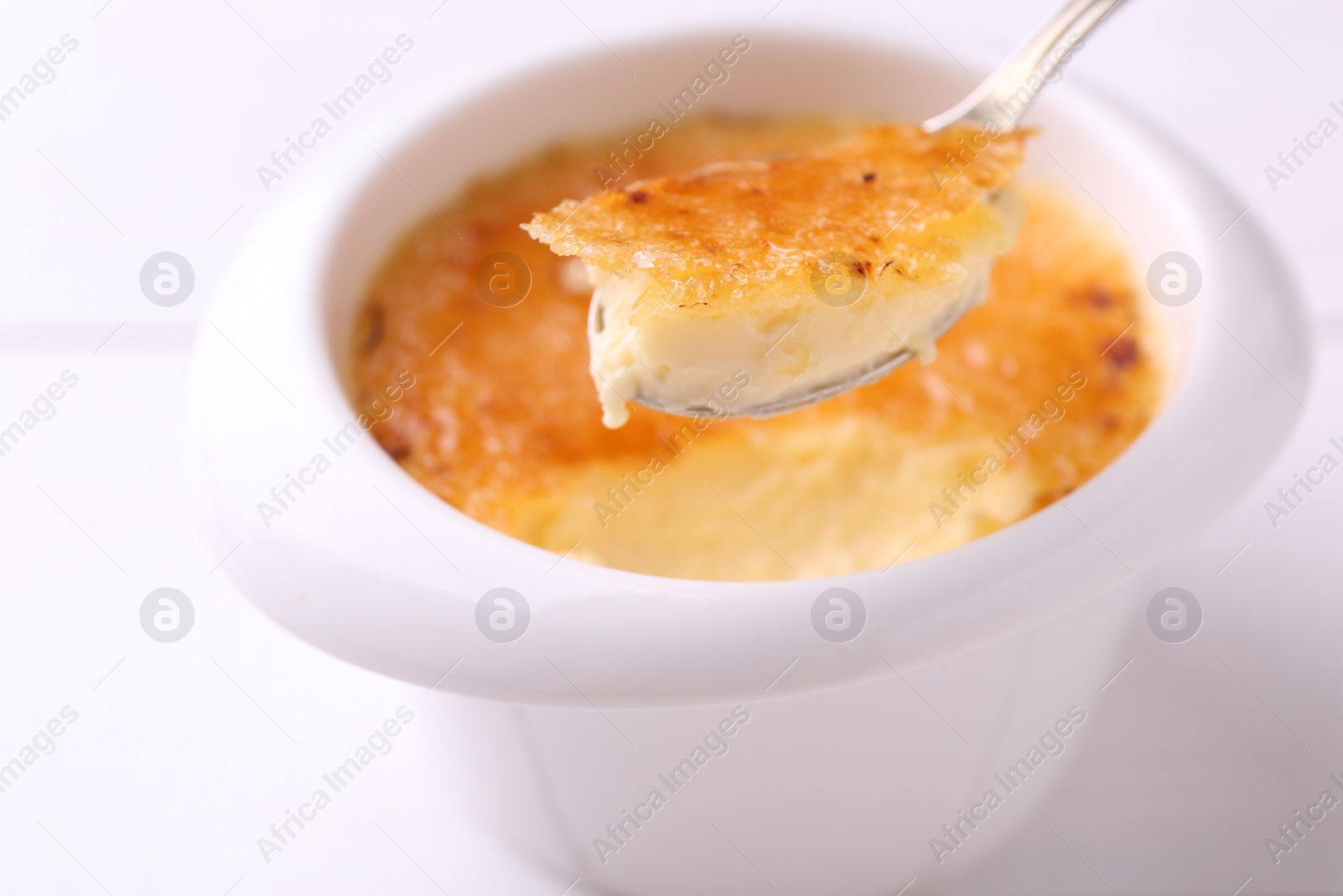 Photo of Taking delicious creme brulee with spoon from bowl at white table, closeup