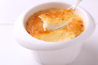 Taking delicious creme brulee with spoon from bowl at white table, closeup