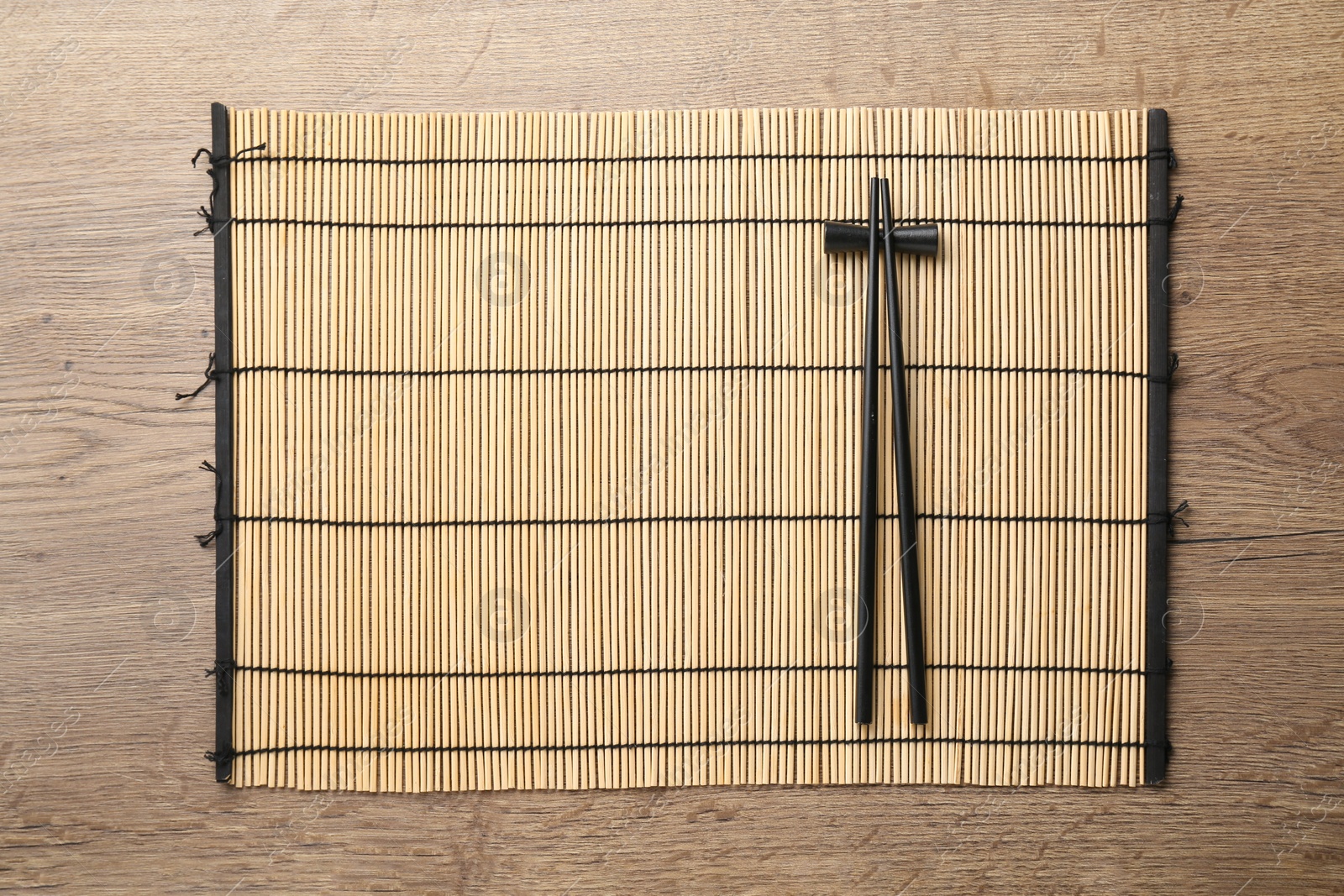 Photo of Bamboo mat with pair of black chopsticks and rest on wooden table, top view