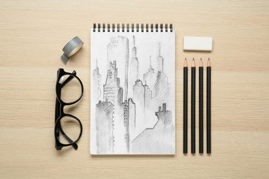Image of Sketch of cityscape in notebook, pencils, eraser and glasses on wooden table, flat lay