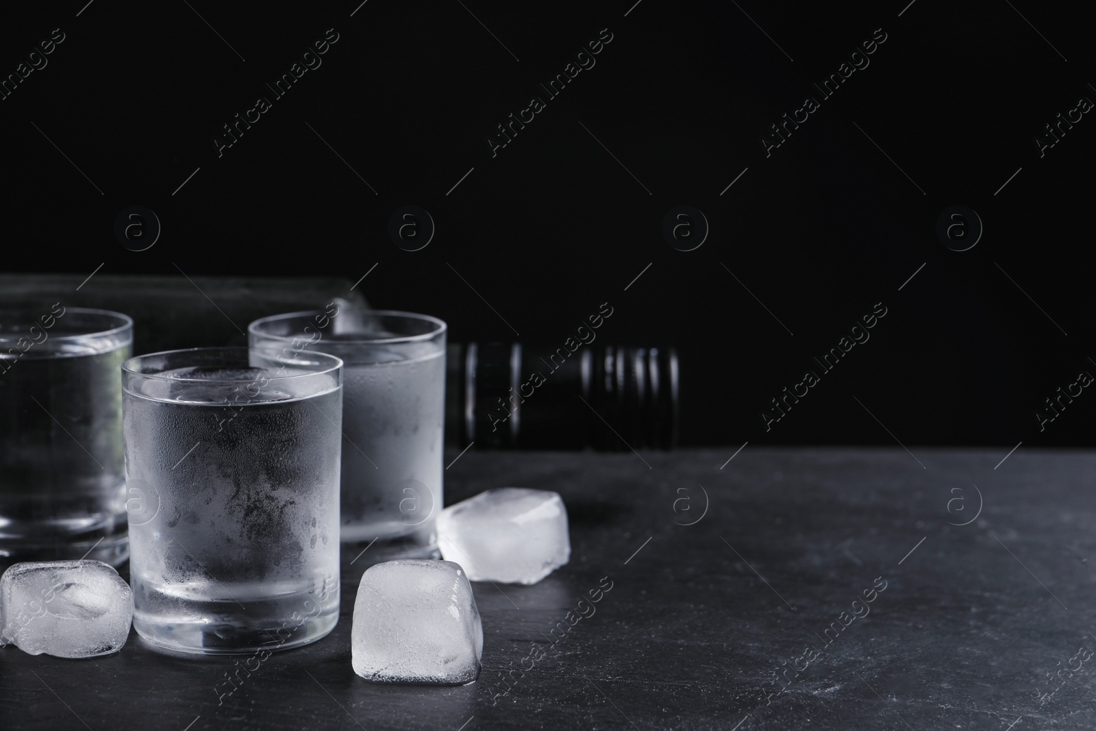 Photo of Bottle of vodka and shot glasses with ice on table against black background. Space for text