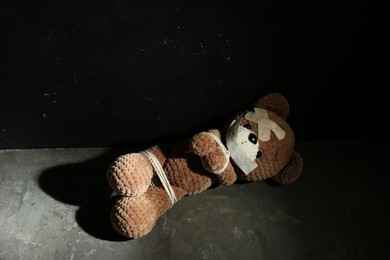 Photo of Stop child abuse. Tied toy bear with taped mouth and patches lying on grey floor against black background. Space for text