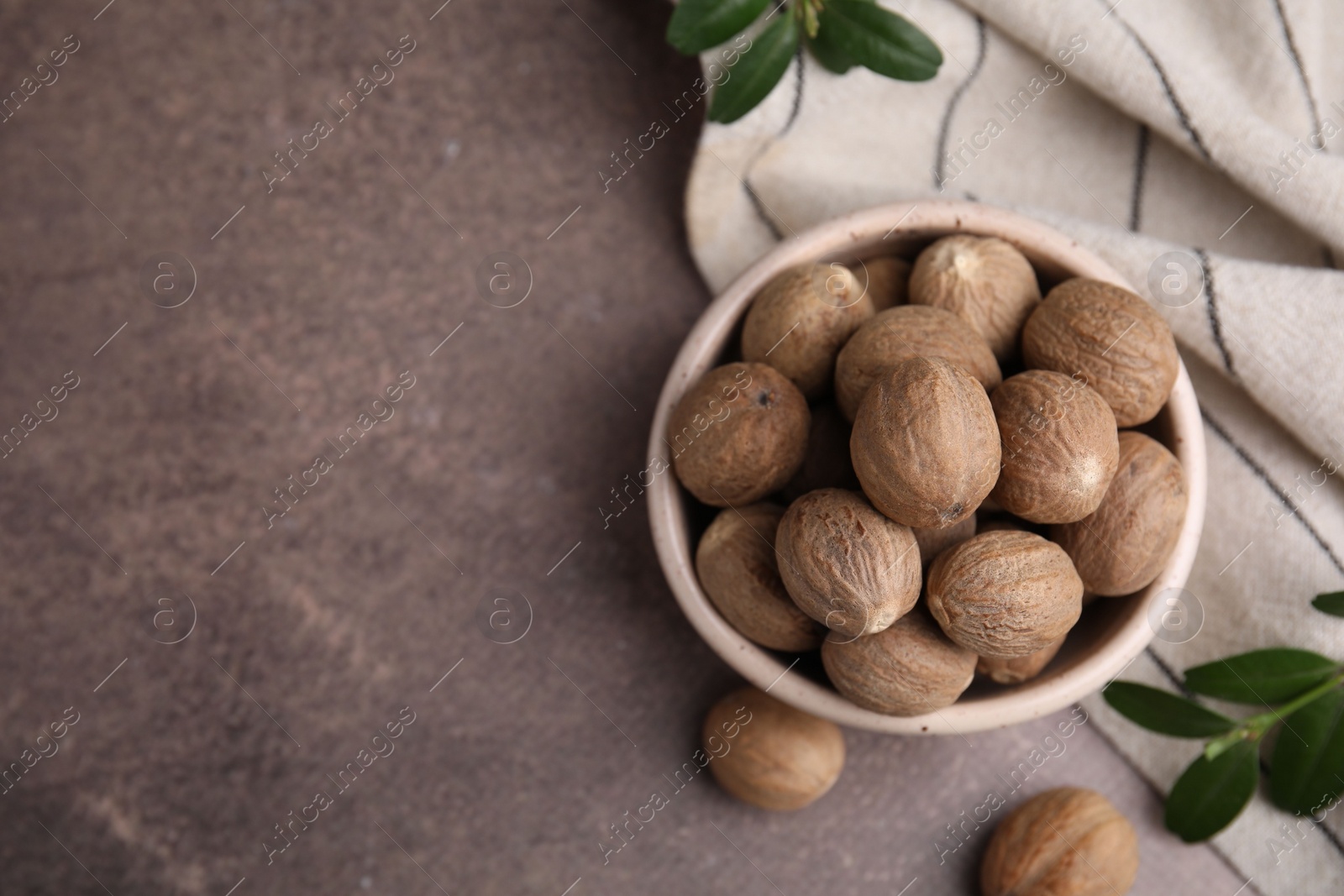 Photo of Whole nutmegs in bowl and green branches on brown table, flat lay. Space for text