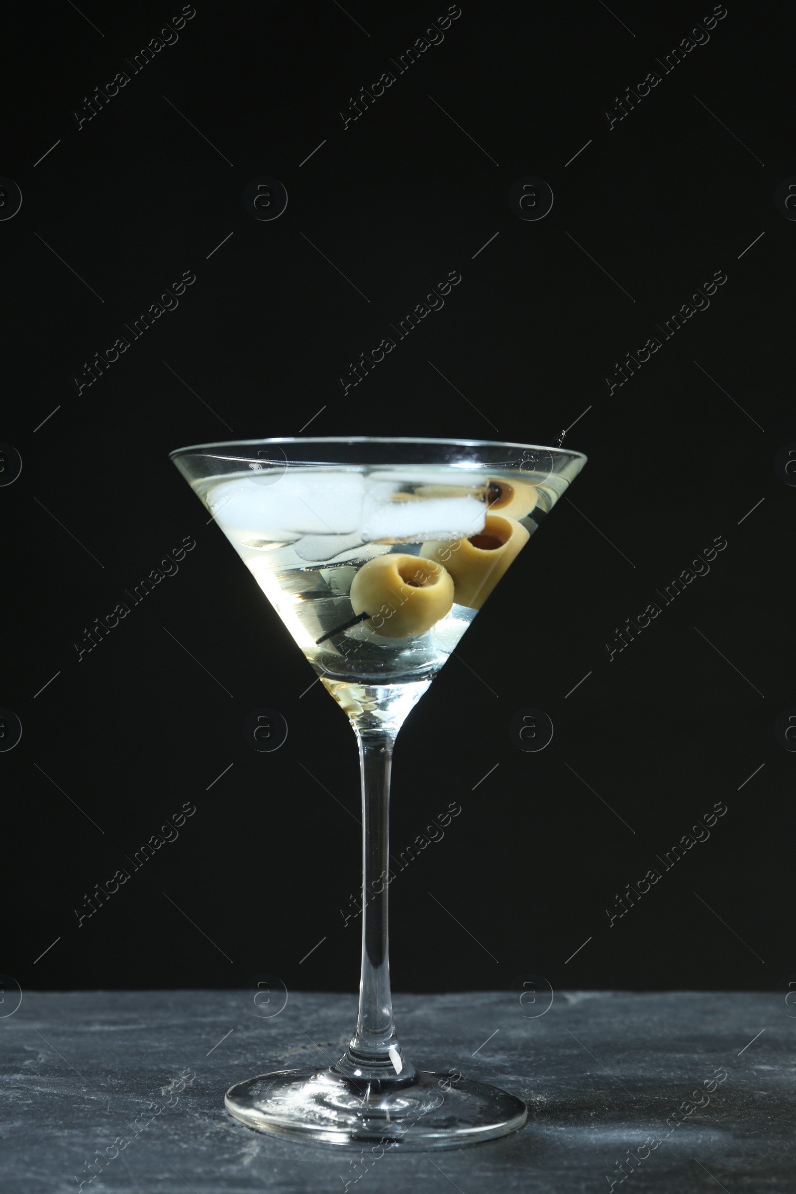 Photo of Martini cocktail with ice and olives on grey table against dark background