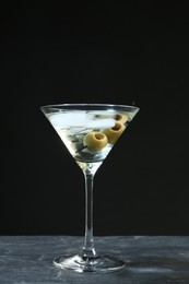 Photo of Martini cocktail with ice and olives on grey table against dark background