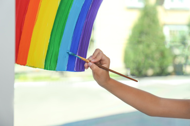 Photo of Little girl drawing rainbow on window indoors, closeup. Stay at home concept