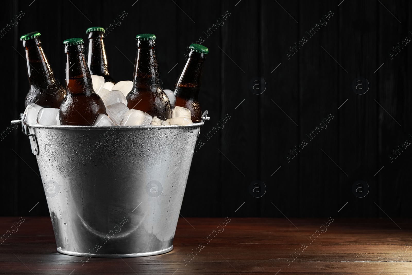 Photo of Metal bucket with bottles of beer and ice cubes on wooden table, space for text